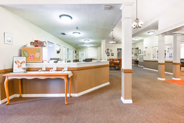 Interior view of a senior living facility reception area with a front desk, a small table with decorations and informational materials, beige walls with white trim, carpeted floor, and columns. In the background, there is a dining area with tables and chairs and a bookshelf along the wall.