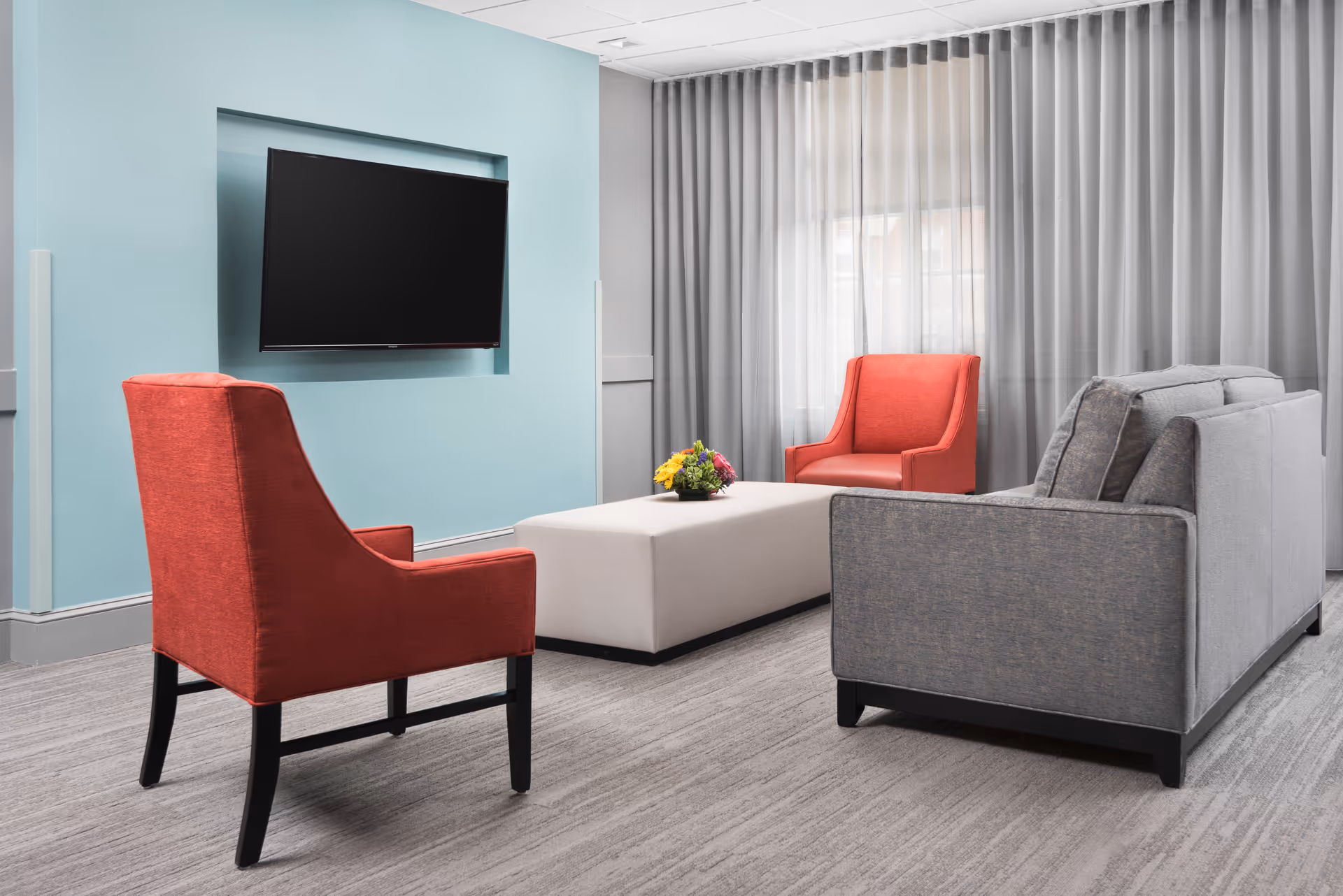 A modern living room area with two red armchairs, a gray sofa, and a white rectangular ottoman with a small flower arrangement on top. A flat-screen TV is mounted on a light blue accent wall, and large windows with sheer gray curtains allow natural light into the room.