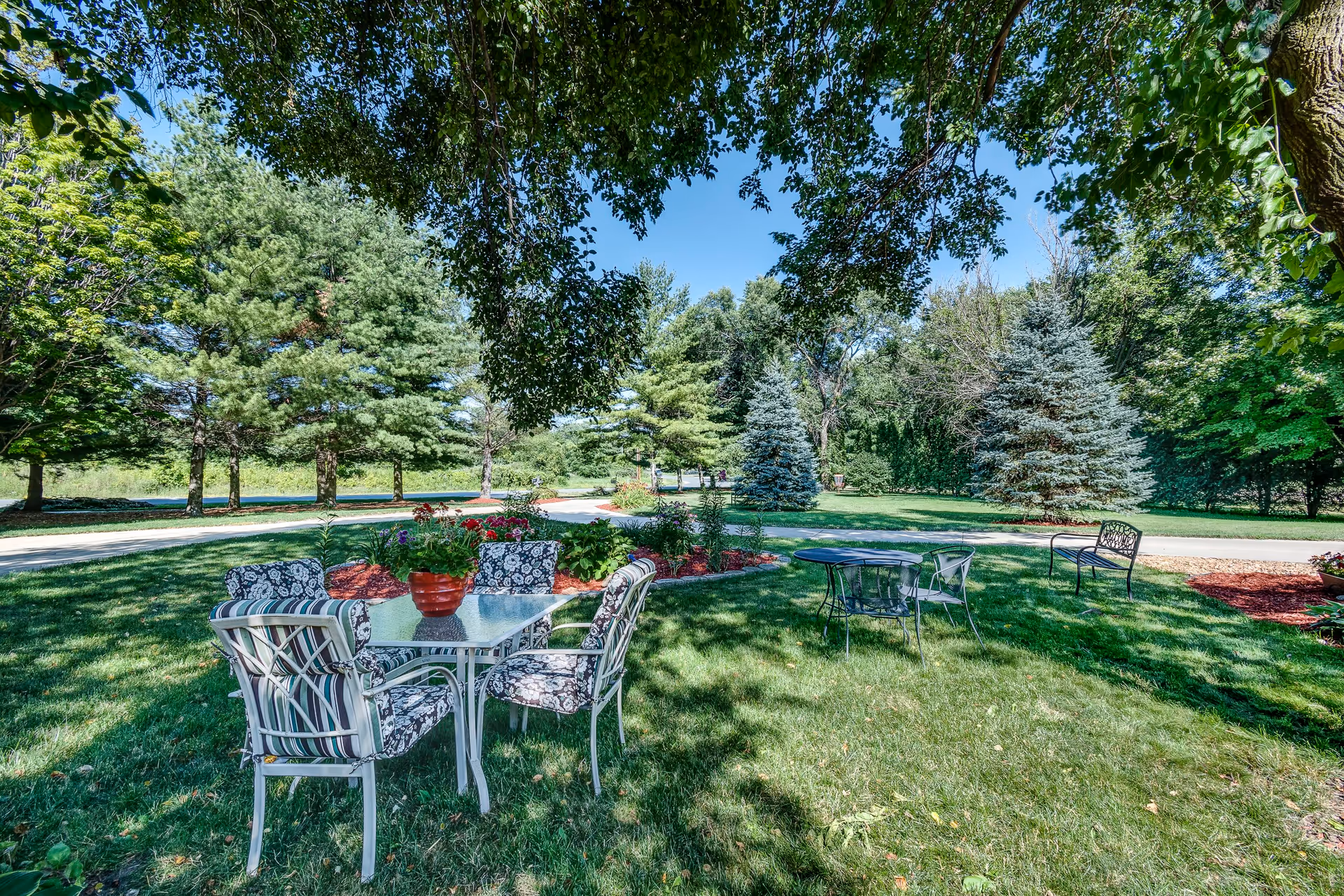 Outdoor garden area with green grass, several trees, and two sets of patio furniture including a glass table with floral cushioned chairs and a smaller round table with metal chairs. There is a flower bed with red mulch and blooming flowers, and a bench is visible in the background under a clear blue sky.