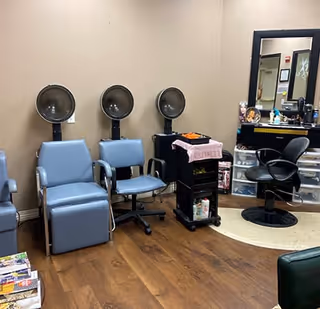 Interior view of a salon area with three blue salon chairs, each positioned under a hair dryer. There is a black rolling cart with hair products and towels, and a black salon chair in front of a large mirror with shelves holding various hair care items. The floor is wooden, and the walls are beige.