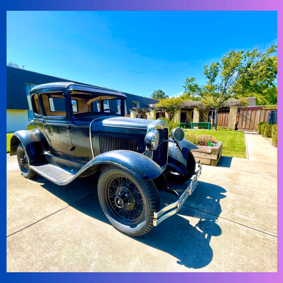 A vintage black car parked on a sunny courtyard in front of a low building with trees, planters, and a walkway.