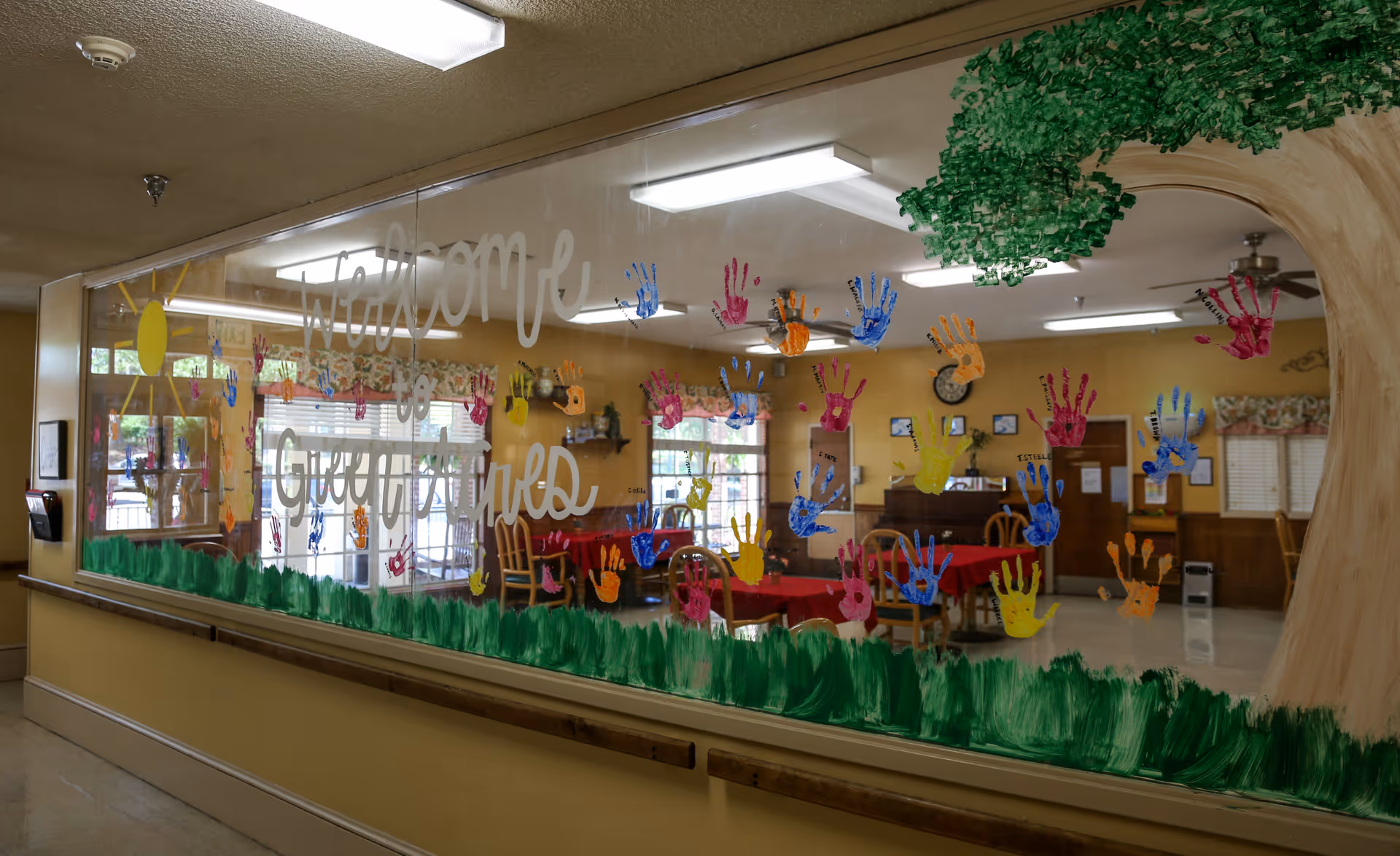 View through a large glass window decorated with colorful handprints and the words 'Welcome to Green Acres' painted on it. Inside, there is a dining area with tables covered in red tablecloths, wooden chairs, and windows with floral curtains. The room has a warm, inviting atmosphere with yellow walls and ceiling lights.