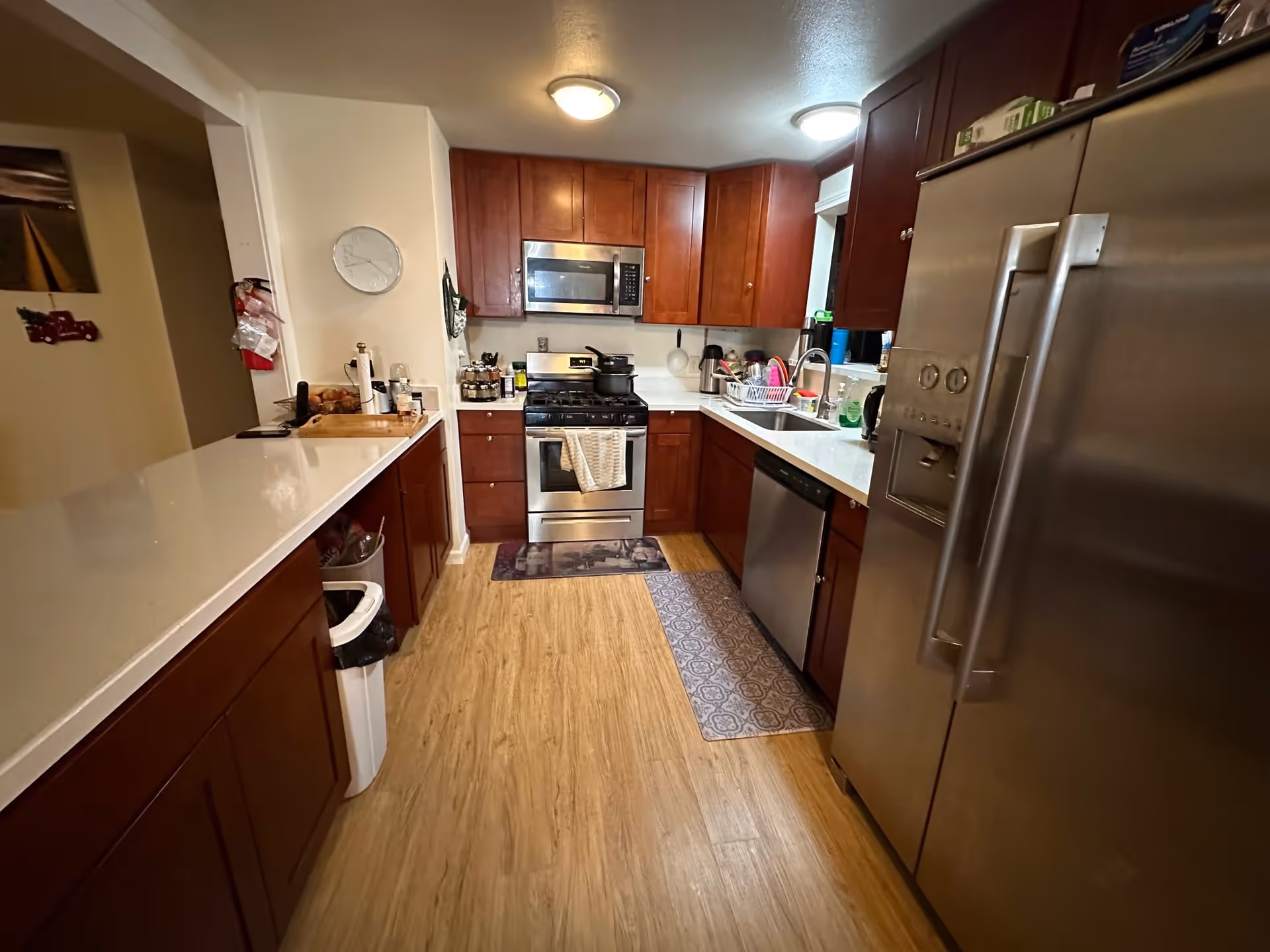 A modern kitchen with stainless steel appliances, wooden cabinets, white countertops, and a refrigerator on the right.