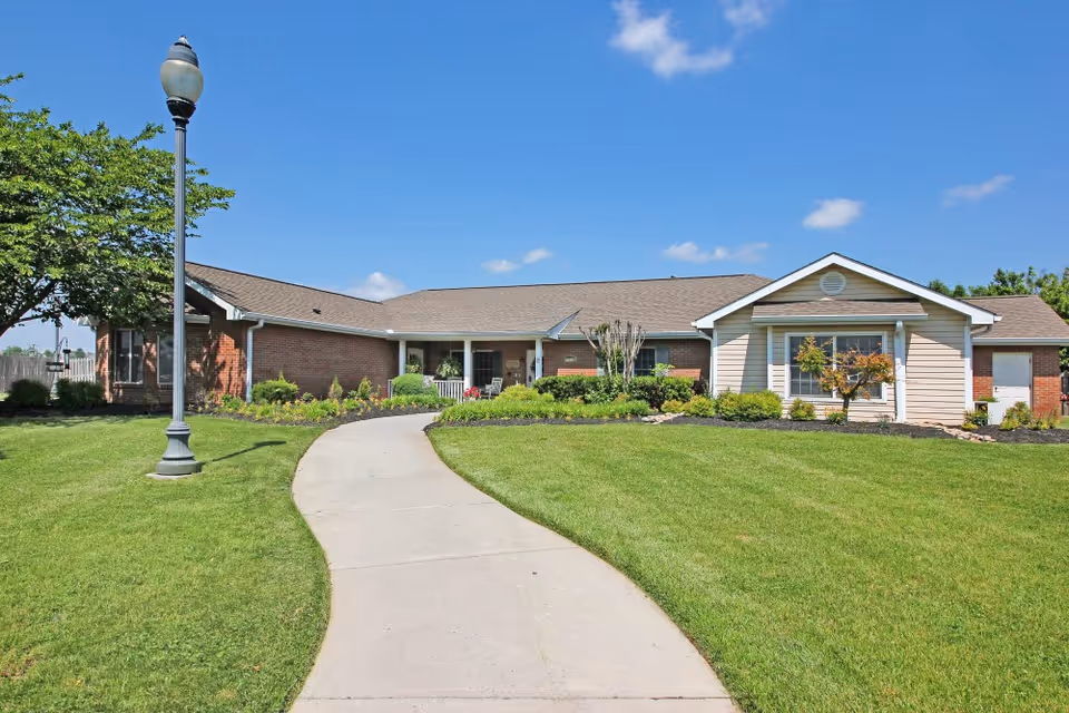 Single-story brick and siding building with a sloped roof, surrounded by a well-maintained lawn and landscaping. A curved concrete pathway leads to the entrance, and a streetlamp stands on the left side near a tree. The sky is clear and blue.