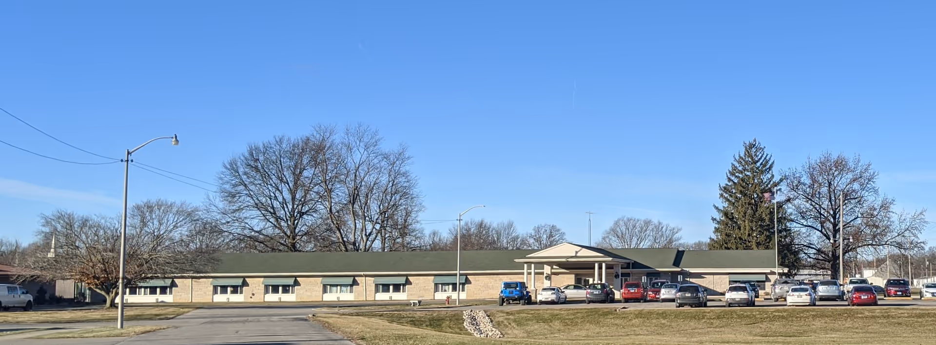 Wide exterior view of a single-story brick building with a green roof, identified as Marshall Rehabilitation and Nursing. The building has multiple windows and a covered entrance with several cars parked in front. Leafless trees and a clear blue sky are visible in the background.