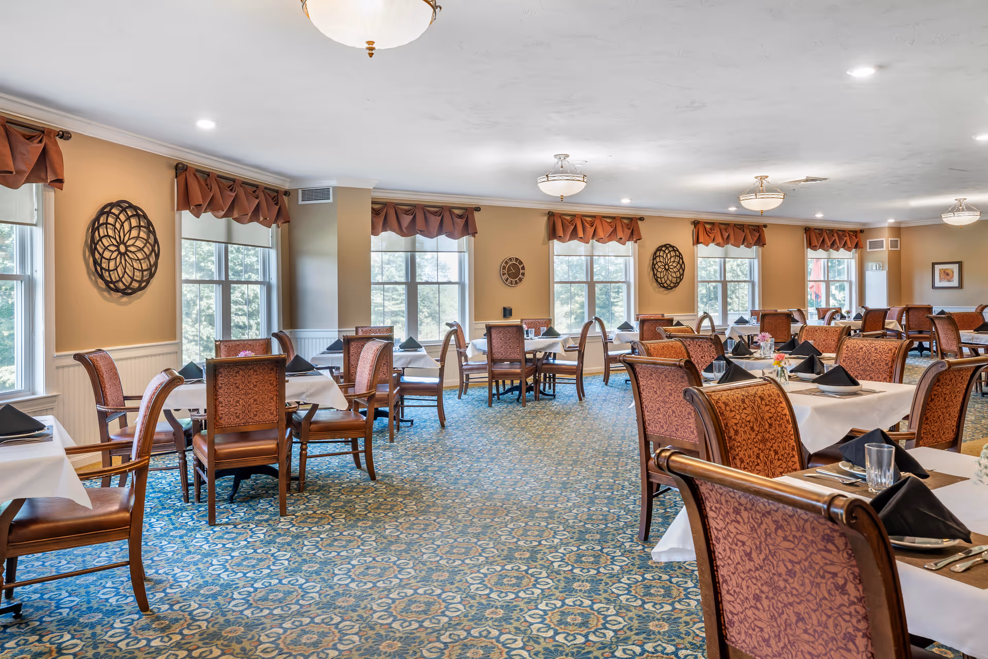 A spacious dining room with multiple tables covered in white tablecloths and set with black napkins, glasses, and silverware. The room features large windows with brown valances, patterned carpet, and decorative wall art. The lighting is provided by ceiling fixtures, creating a bright and welcoming atmosphere.