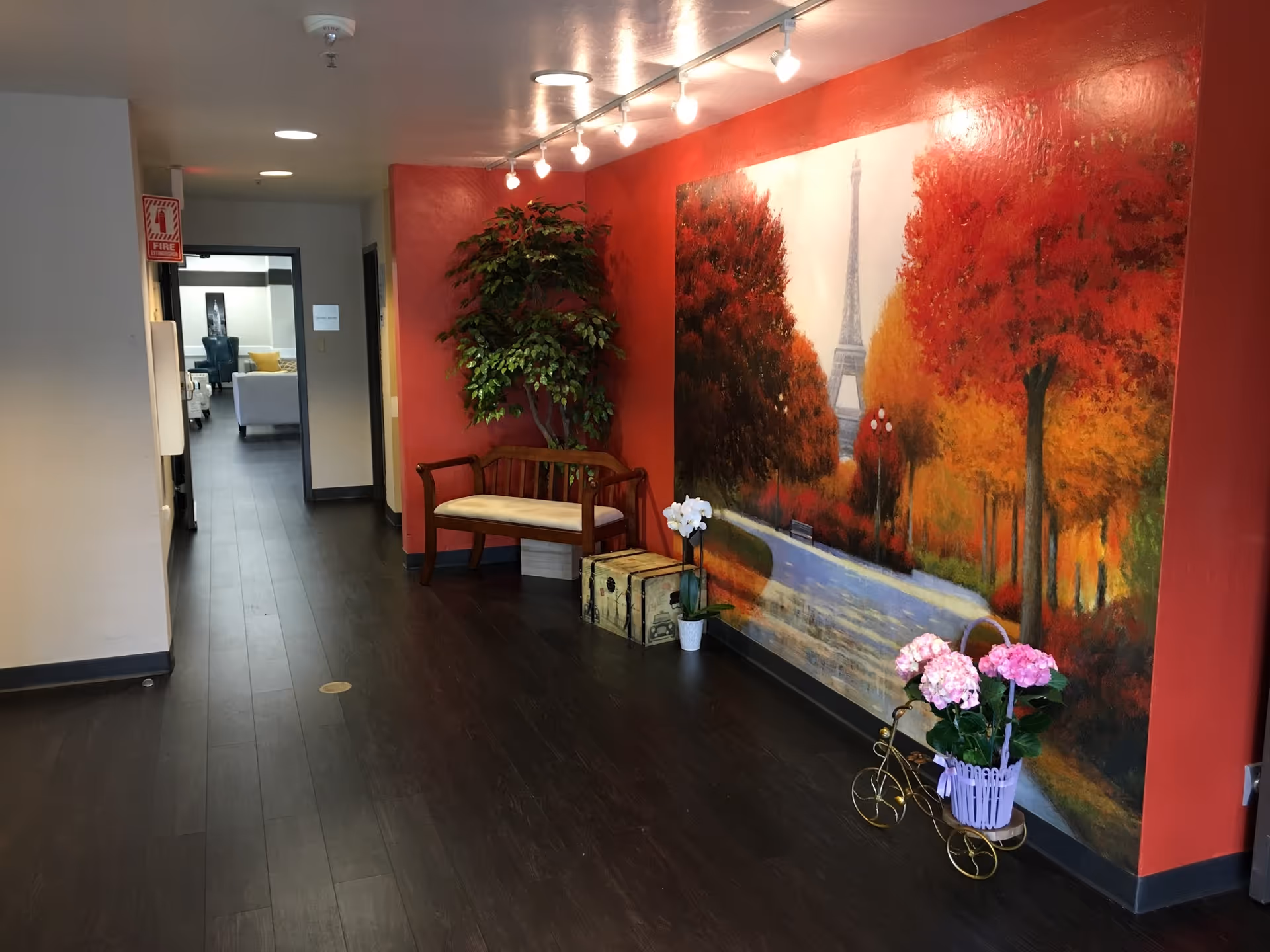 Interior hallway with dark wood floor, a red accent wall mural of the Eiffel Tower, a bench, plants, and floral decorations.