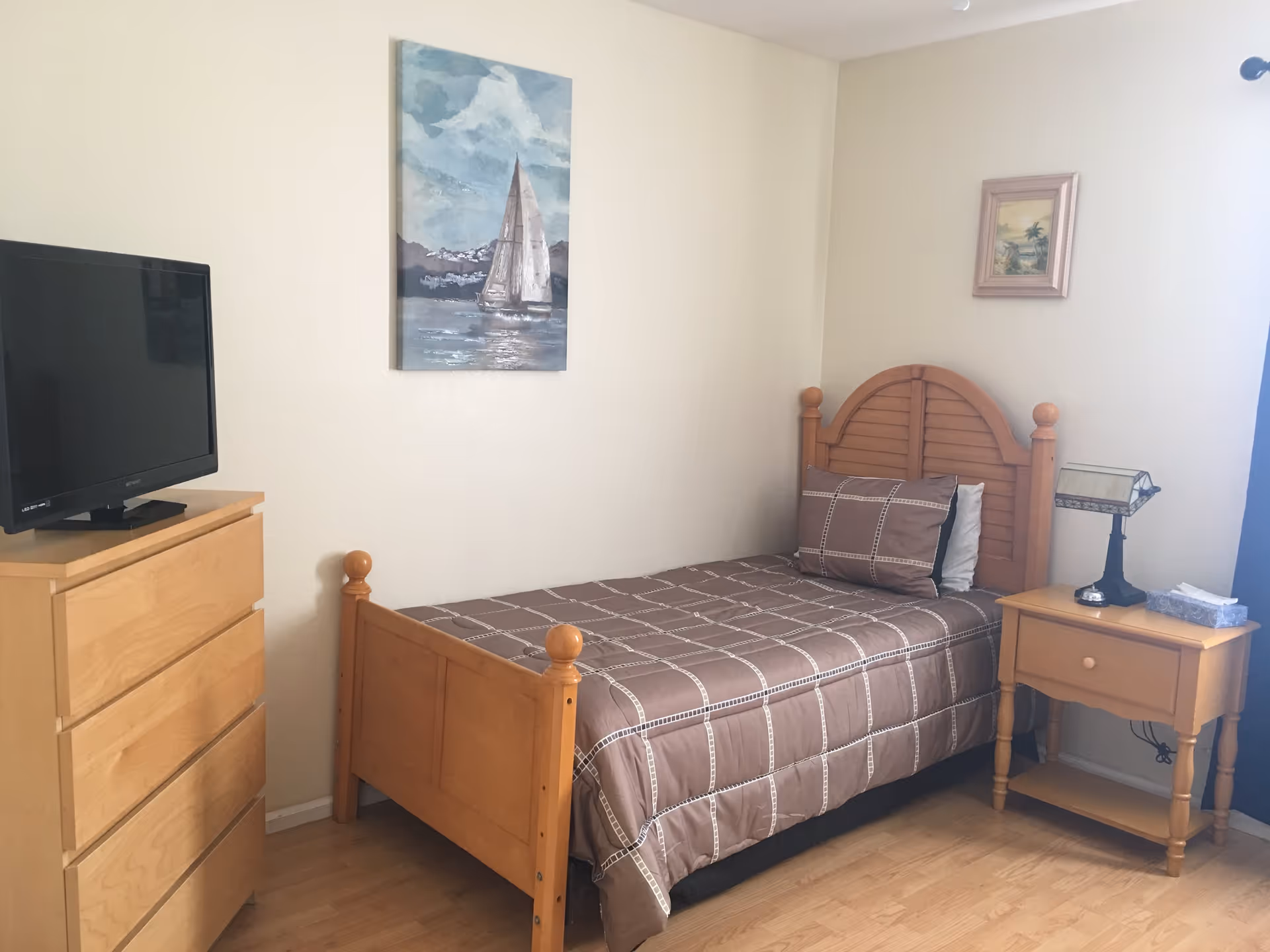 A small bedroom with a single wooden bed covered with a brown checkered bedspread and matching pillow. Next to the bed is a wooden nightstand with a decorative lamp and a box of tissues. On the opposite side, there is a wooden dresser with a flat-screen TV on top. The walls are light-colored and decorated with two framed paintings, one of a sailboat and another of a tropical scene. The floor is wooden.