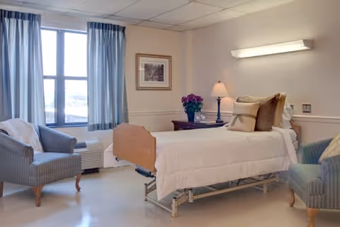 A bright and clean patient room with a hospital bed, two upholstered armchairs, a nightstand with a lamp and a vase of flowers, a framed picture on the wall, and large windows with blue curtains letting in natural light.
