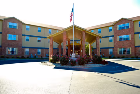 Front exterior view of a three-story senior living facility building with a covered entrance supported by brick columns, landscaped with bushes and flowers, and an American flag flying on a pole above the entrance.