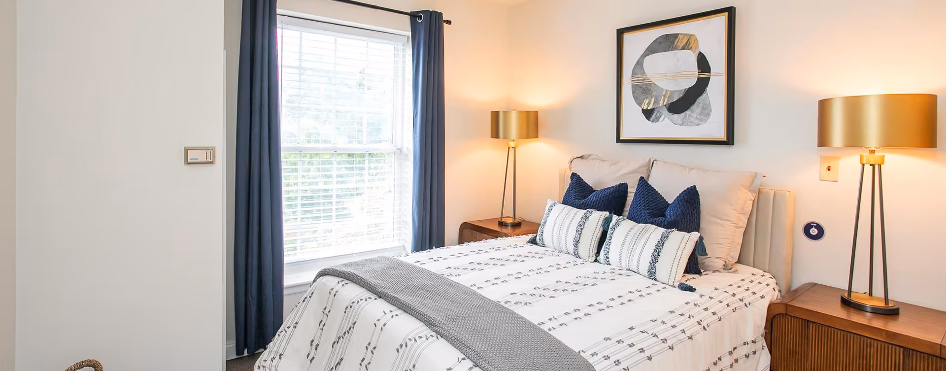 A neatly made bed with white and patterned bedding, adorned with blue and white pillows, positioned between two wooden nightstands each holding a modern gold lamp. A large window with white blinds and dark blue curtains is on the left side, and a framed abstract artwork hangs on the wall above the bed.