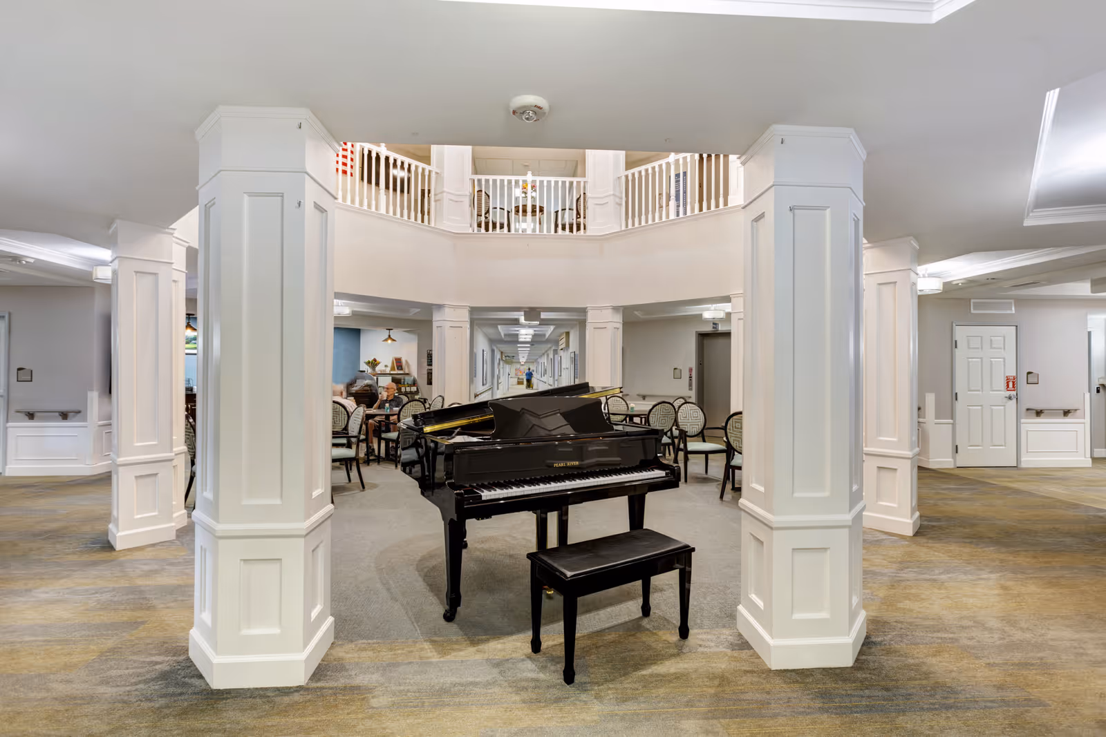 Interior view of a senior living facility common area featuring a black grand piano with a matching bench in the center, surrounded by white pillars and seating areas with chairs and tables. The space has a carpeted floor and a second-floor balcony with white railings overlooking the area.