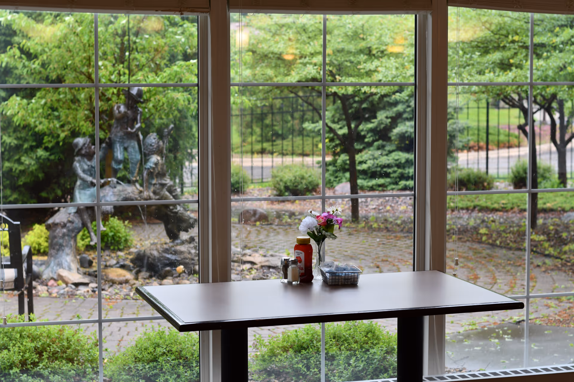 A table with a small flower vase, ketchup bottle, salt and pepper shakers, and a basket with packets is positioned in front of large windows. Outside the windows, there is a garden area with green bushes, trees, a paved walkway, and a bronze statue of children playing on a tree branch.