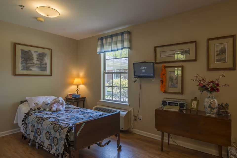 A cozy bedroom in a senior living facility featuring a single bed with a patterned blanket and several stuffed animals on it. There is a small wooden nightstand with a lamp and a tissue box next to the bed. A window with a blue and white valance lets in natural light. On the opposite wall, a small flat-screen TV is mounted above a wooden table decorated with framed pictures, a vase with flowers, and a radio. The walls are adorned with framed landscape paintings, and the floor is wooden.