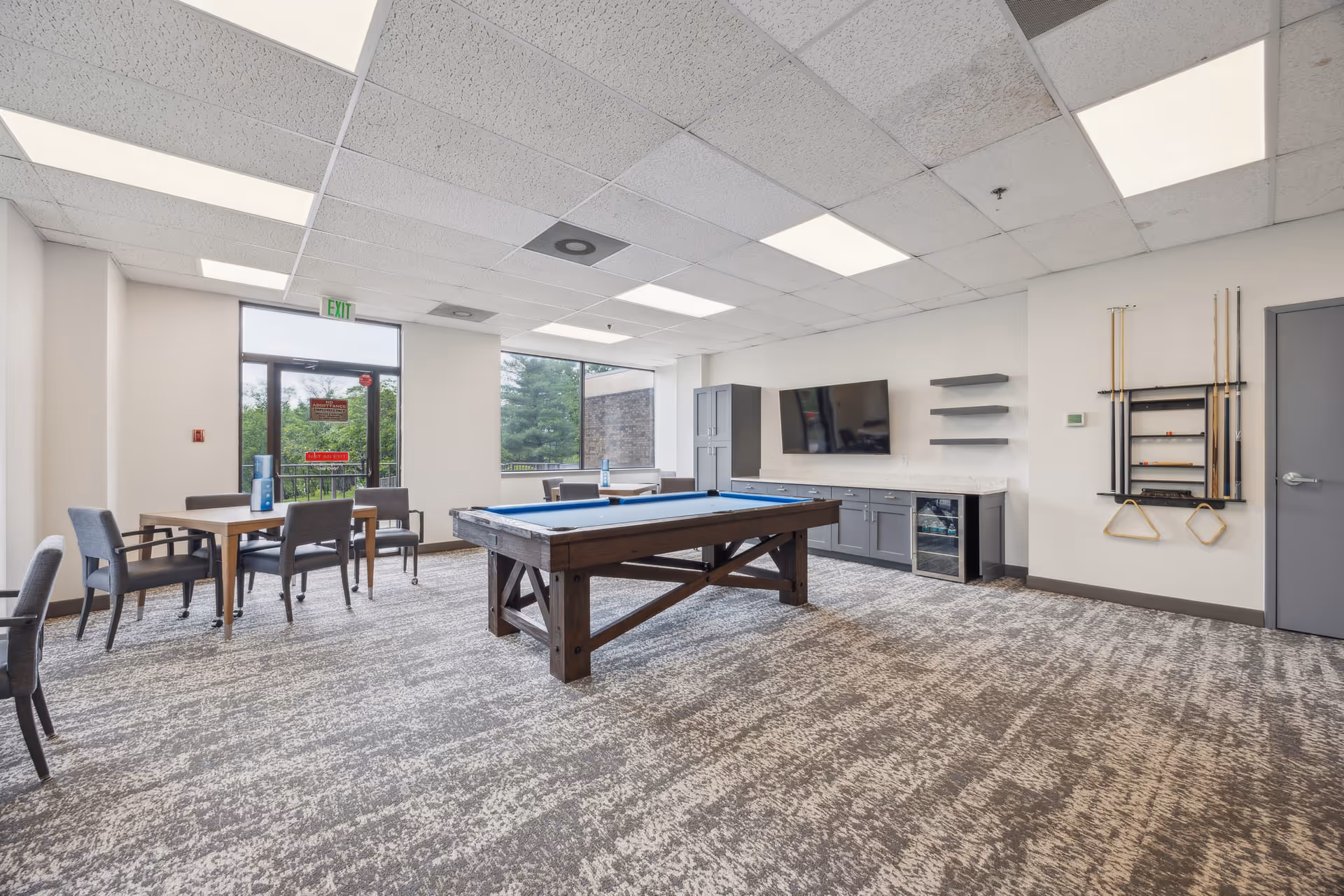 Bright interior game room with a pool table, tables and chairs, wall-mounted TV, and cue rack.
