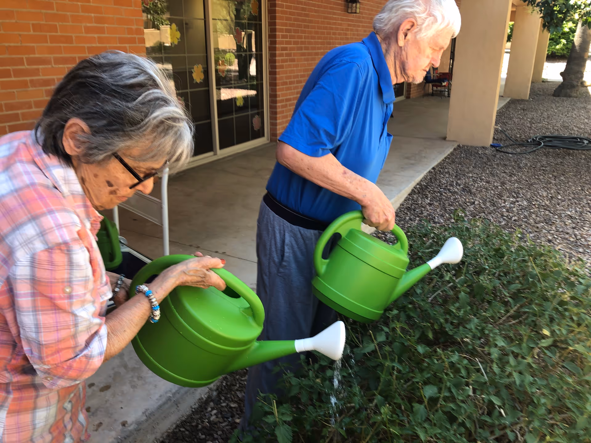Two elderly individuals watering plants with green watering cans outside a brick building with a covered walkway.