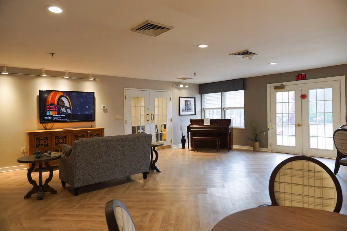 A spacious living room area in a senior living facility with a gray couch facing a wall-mounted flat-screen TV. There is a wooden piano against the far wall near two windows with blinds, and a framed picture hanging above it. Double glass doors and a single glass door with an exit sign above are visible, along with a round table and chairs in the foreground. The room has wooden flooring and neutral-colored walls.