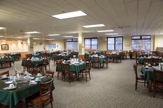 A spacious dining room with multiple round tables covered with green tablecloths, each set with plates, cups, and silverware. The room has beige walls, large windows letting in natural light, and a carpeted floor. Ceiling lights illuminate the area evenly.