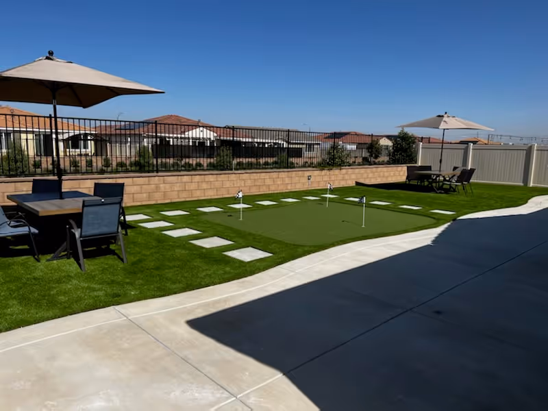 Outdoor patio area with artificial grass, a small putting green with four golf holes, two seating areas each with a table, four chairs, and a large umbrella. The area is enclosed by a low brick wall and a metal fence, with houses visible in the background under a clear blue sky.
