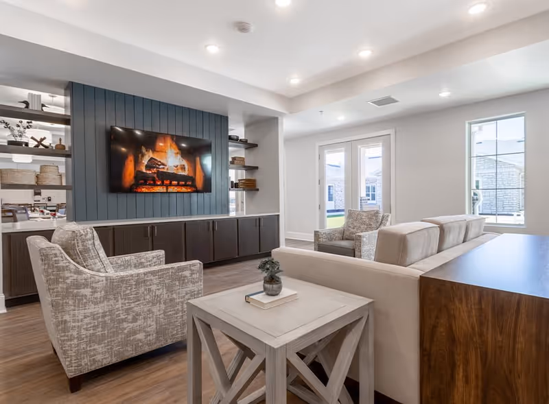 A modern living room with a beige sofa, two patterned armchairs, and a light wooden coffee table with a small plant and book on it. A flat-screen TV mounted on a blue paneled wall displays a burning fireplace. The room has recessed lighting, wooden flooring, and large windows with a view outside.