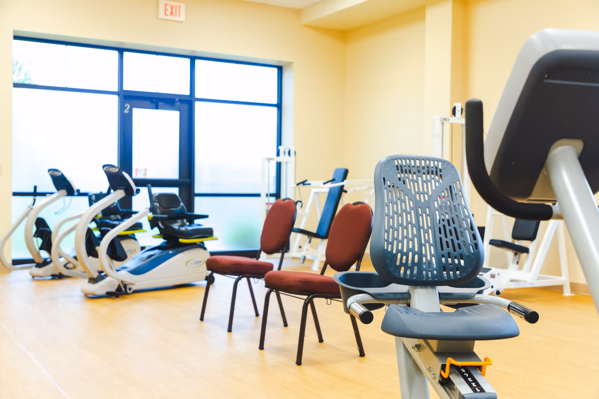 A bright exercise room with several pieces of fitness equipment including recumbent bikes and weight machines, along with two red cushioned chairs. Large windows and a door with frosted glass allow natural light to fill the space.