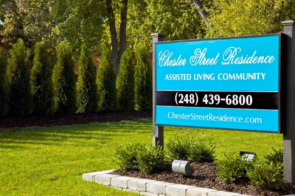 Outdoor view of a sign for Chester Street Residence Assisted Living Community, placed on a grassy area with bushes and trees in the background.