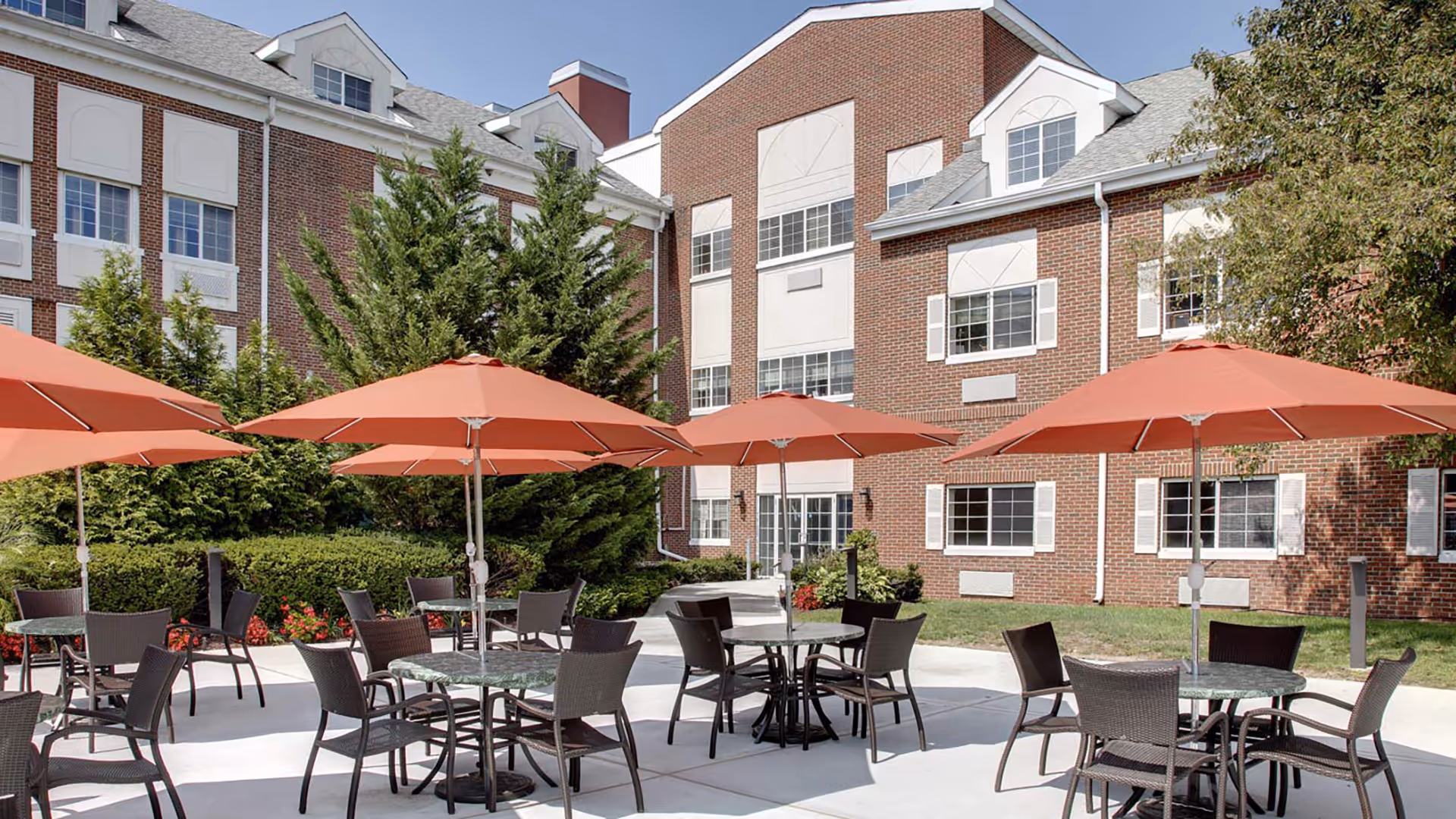 Outdoor patio area at a senior living facility with several round tables, each shaded by large orange umbrellas. Each table is surrounded by dark wicker chairs. The patio is adjacent to a multi-story brick building with white-trimmed windows and surrounded by greenery including trees and bushes.