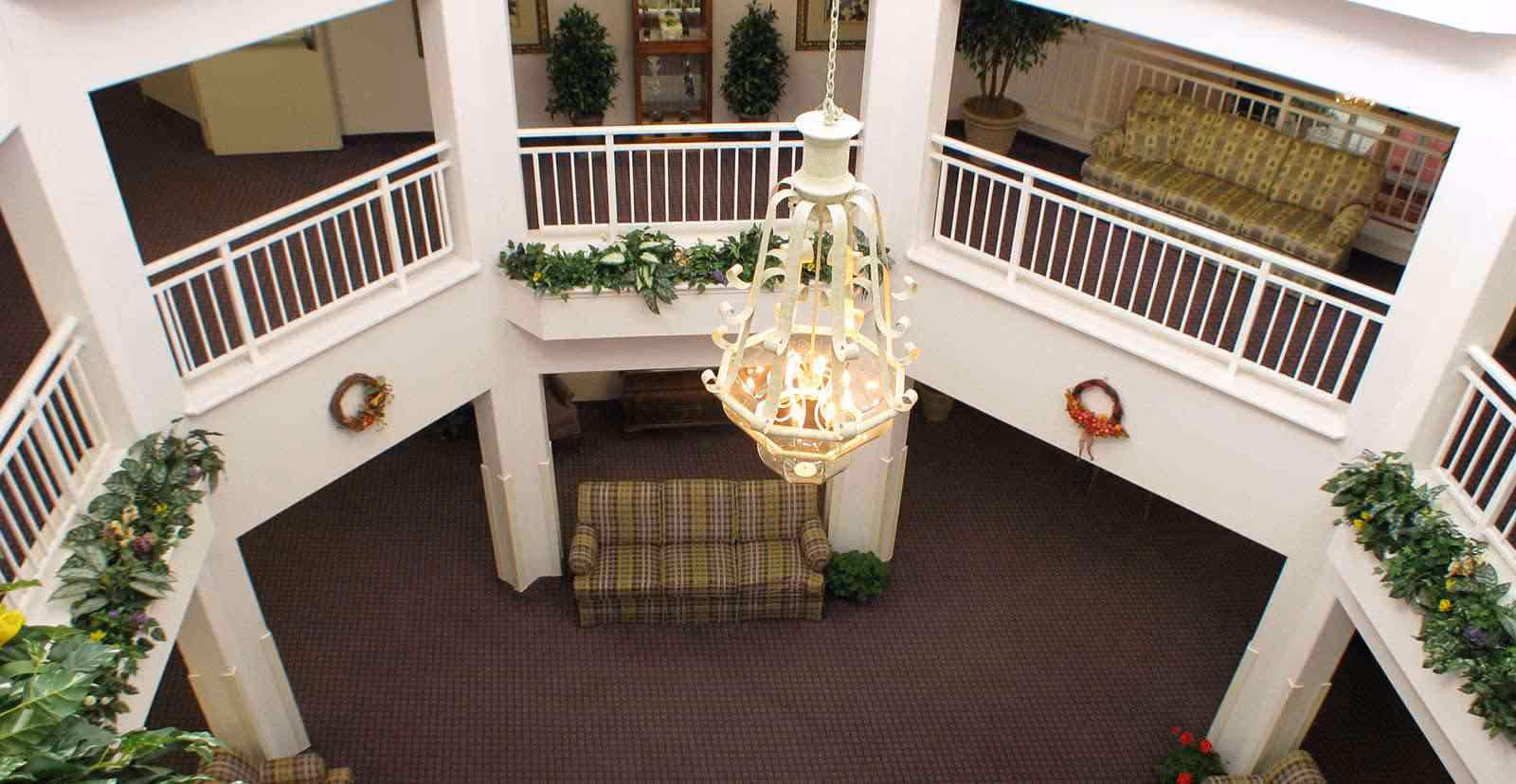 Interior view of a senior living facility atrium with a large white chandelier hanging from the ceiling. The space features a two-story open area with white railings, green plants along the edges, and plaid upholstered sofas on both levels. The carpet is dark and patterned, and there are decorative wreaths on the walls.
