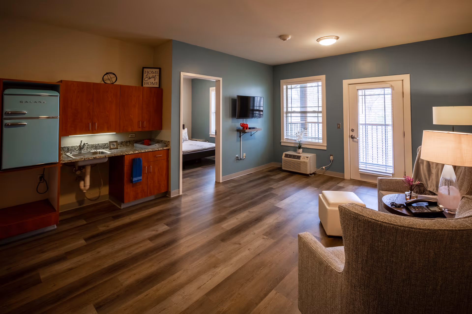 Interior view of a senior living facility apartment with a small kitchenette featuring a retro-style blue refrigerator, wooden cabinets, and a granite countertop with a sink. The room has wood flooring and blue walls. A doorway leads to a bedroom with a bed. The living area includes a wall-mounted TV, a window, a door with blinds leading outside, a beige armchair, a small round table with a lamp, and a white ottoman.