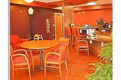 Interior view of a common area in Thornapple Manor featuring a round wooden table with four red cushioned chairs, a tall brick column, a counter with bar stools, warm orange walls, and a floor lamp. There are plants visible in the foreground.