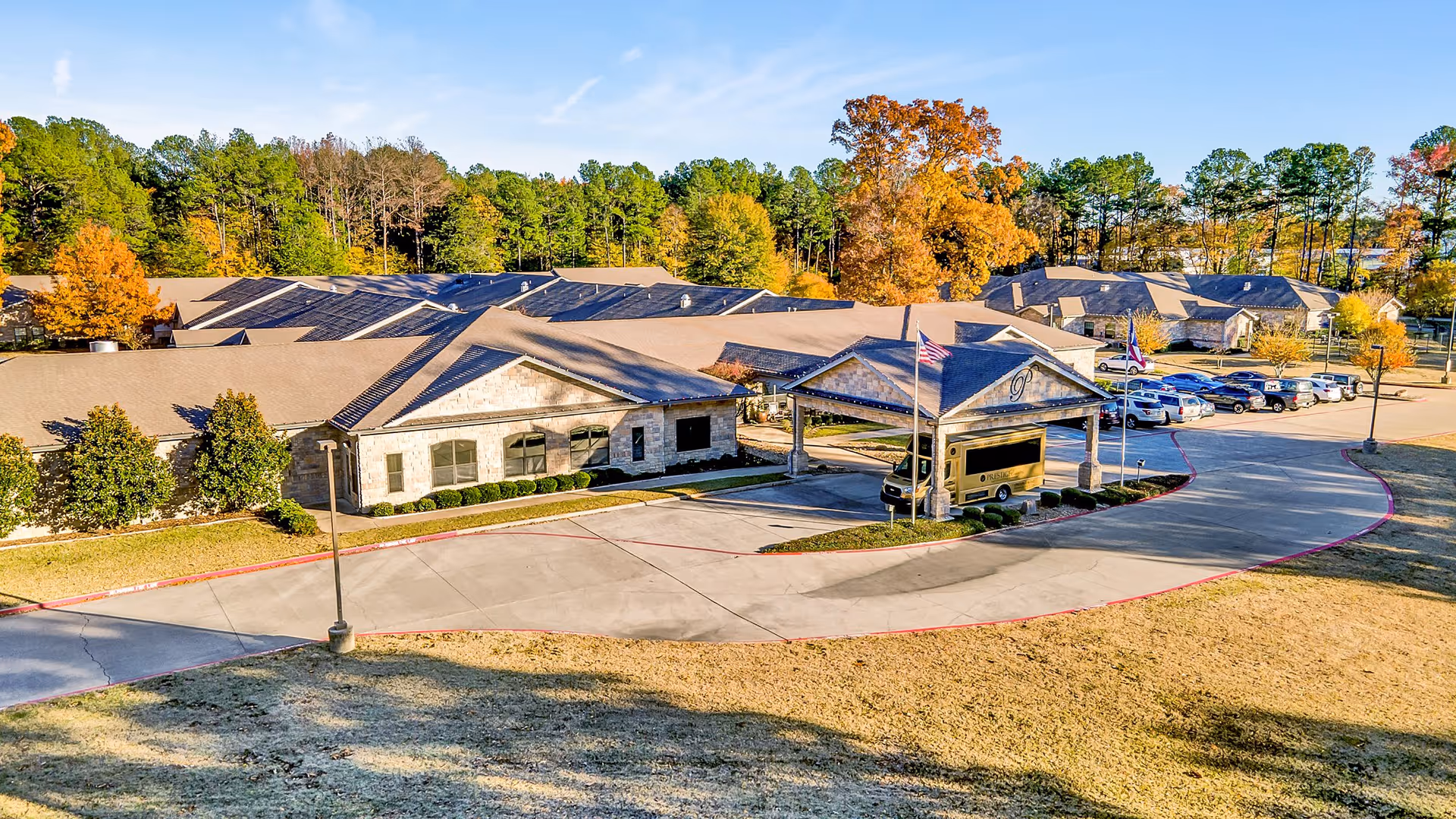 Aerial view of Prestige Estates Assisted Living & Memory Care facility showing a single-story building with a covered entrance, a parking lot with several cars, surrounded by trees with autumn foliage under a clear blue sky.