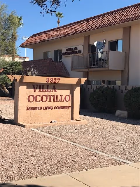Exterior view of Villa Ocotillo Assisted Living Community building with a beige sign in front displaying the name and address 3327. The building has a tiled roof, balcony with a satellite dish, and desert landscaping with gravel and bushes.