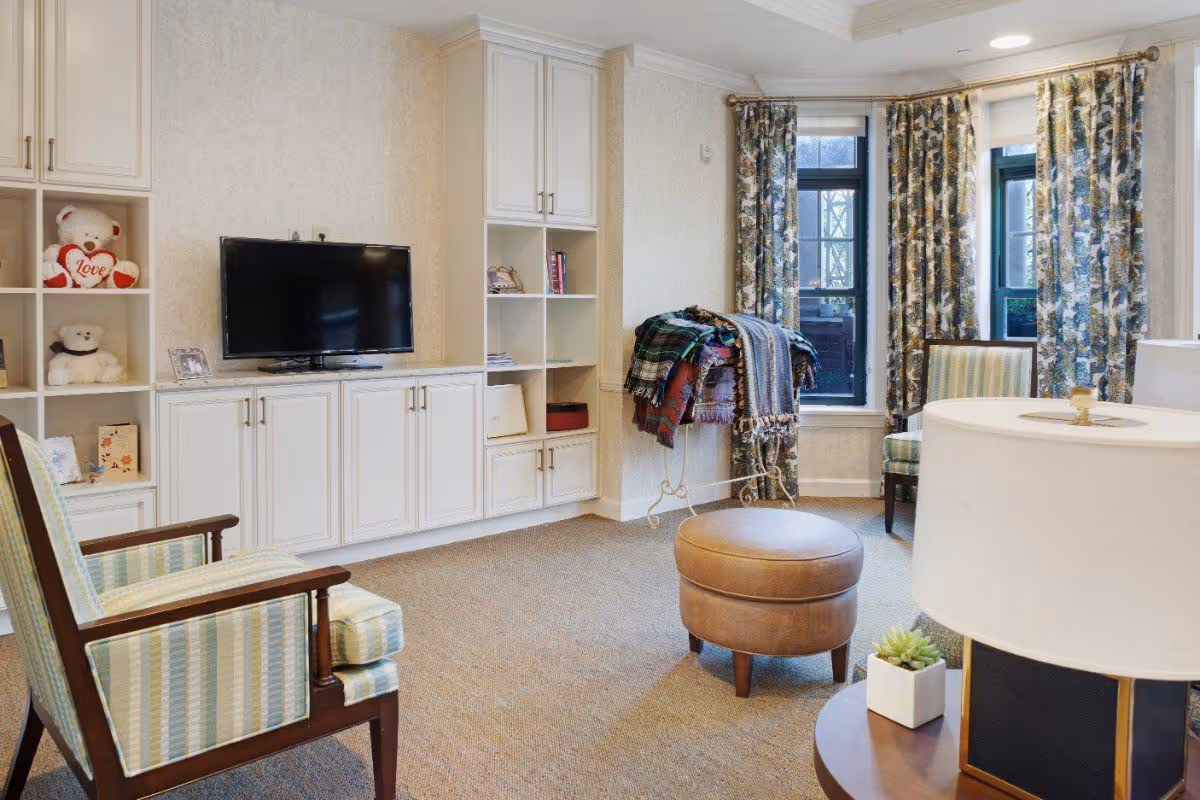 A cozy living room with two striped armchairs, a round leather ottoman, a flat-screen TV mounted on a white built-in cabinet with shelves displaying teddy bears and books, floral curtains on windows, and a small table with a lamp and a succulent plant.