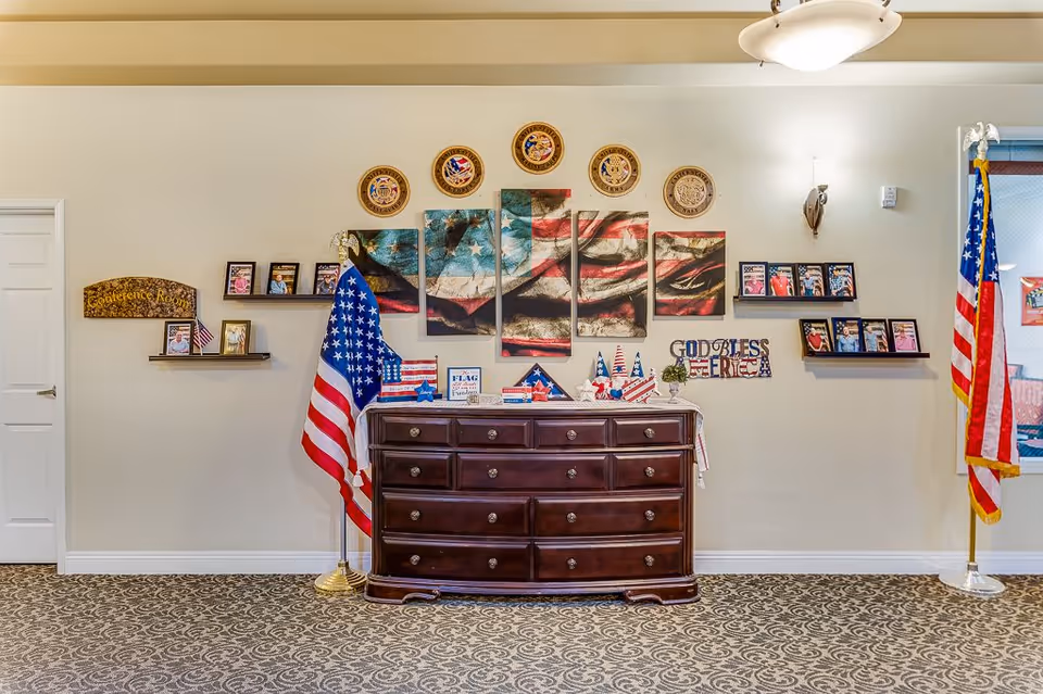 Interior wall decorated with patriotic American flags and memorabilia, including a large multi-panel artwork of the American flag, several smaller flags, plaques, and framed photos. A dark wooden dresser is placed against the wall, flanked by two American flags on stands. The wall also features a sign that reads 'Conference Room' and another that says 'God Bless America'.