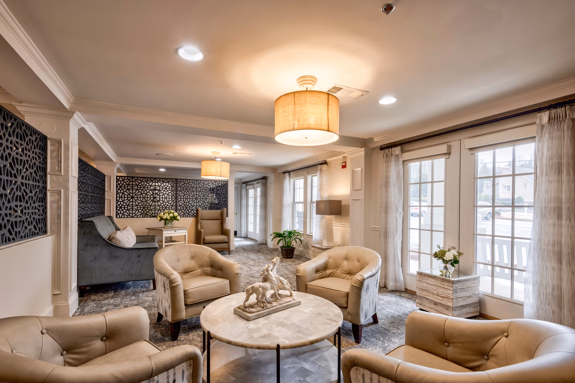 A cozy and well-lit living room area with four beige armchairs arranged around a round marble coffee table that has a decorative horse sculpture on it. The room features large windows with sheer curtains, two ceiling light fixtures with beige lampshades, a gray sofa, a beige armchair, a side table with a vase of flowers, and decorative wall panels.