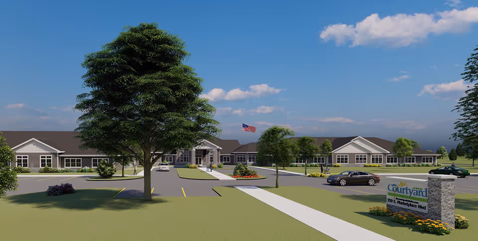 Exterior view of The Courtyard at Delta senior living facility showing a single-story building with a dark roof, multiple windows, and an American flag flying in front. There are trees, landscaped areas, parked cars, and a stone sign with the facility name and address near the parking lot under a partly cloudy blue sky.