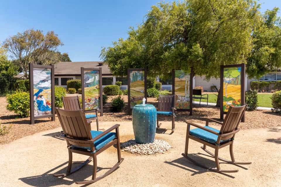 Four wooden rocking chairs with blue cushions arranged around a small blue fountain in a sunny outdoor courtyard with decorative painted panels and trees.