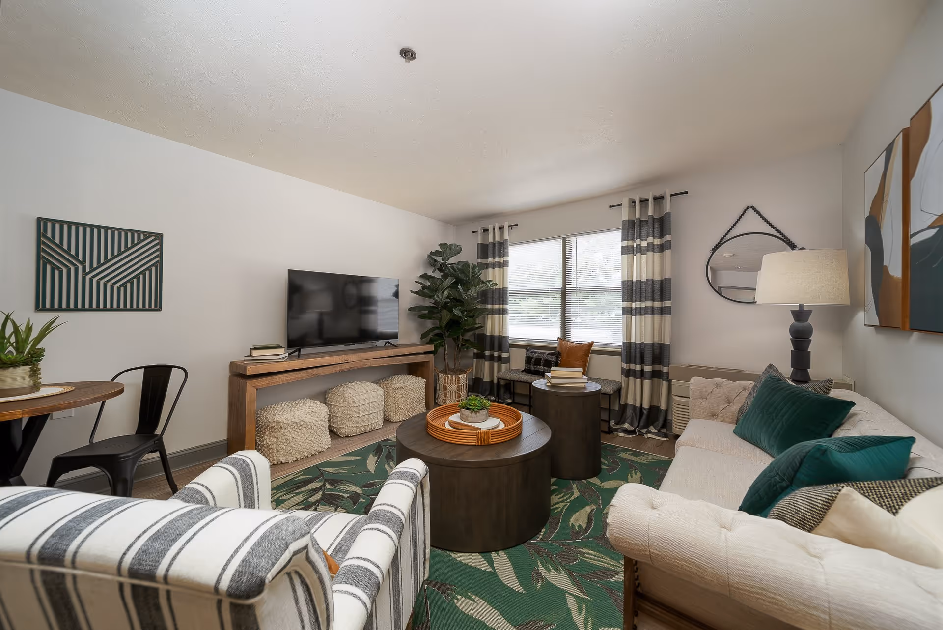 A cozy living room with a beige tufted sofa adorned with green and beige pillows, a striped armchair, and a round wooden coffee table with a decorative tray and plant. There is a flat-screen TV on a wooden console table, two poufs underneath, a large window with striped curtains, a floor lamp, and wall art including a geometric piece and a round mirror.