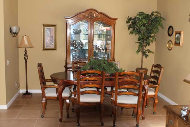 A dining room with a wooden dining table surrounded by six matching wooden chairs with cushioned seats. Behind the table is a wooden china cabinet displaying glassware and decorative items. The room has beige walls with framed artwork, a floor lamp, a wall sconce, and a potted plant in the corner.