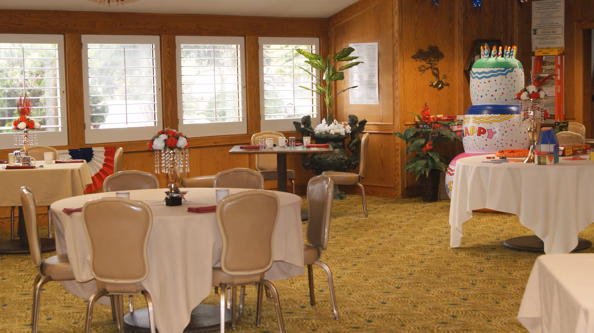 A dining room with round tables covered in white tablecloths and beige chairs around them. Each table has a centerpiece with red and white flowers. The room has wood-paneled walls and windows with white shutters. There is a large, colorful birthday cake decoration on a table to the right, along with some board games and plants in the background.