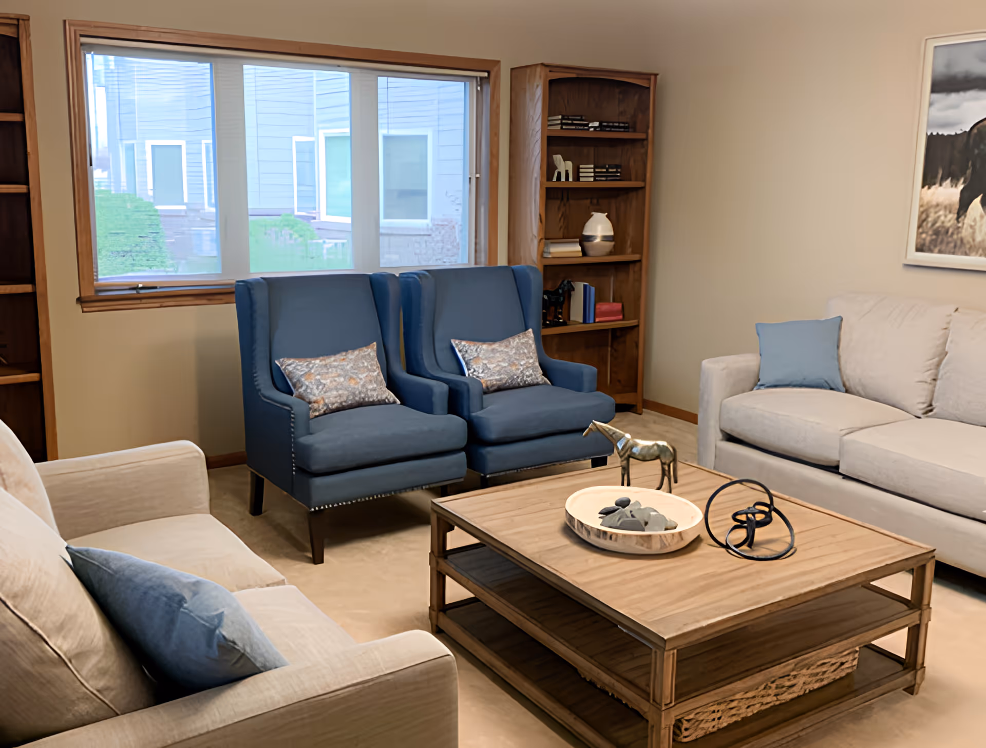 Comfortable living room with two blue armchairs, beige sofas, a wooden coffee table with decorative items, bookshelves and a window.