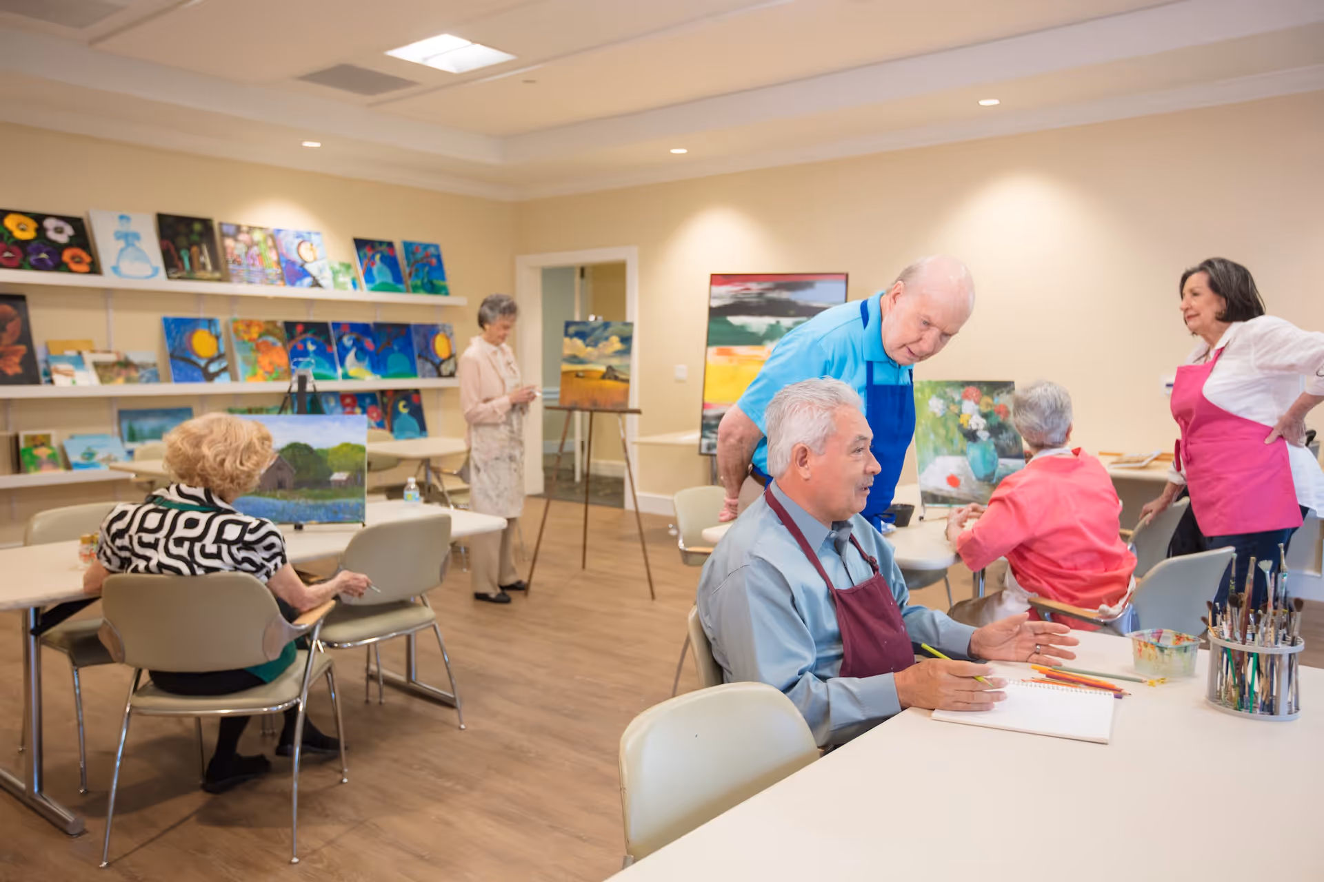 A group of elderly people participating in a painting class in a well-lit room with wooden floors and beige walls. Several paintings are displayed on shelves and easels around the room. Some participants are seated at tables working on their art, while others are standing and interacting. Two women wearing pink aprons appear to be assisting or instructing the group.