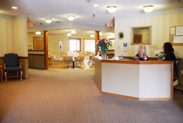 Interior view of a senior living facility reception area with a front desk where two women are conversing. Behind the desk is a mirror and bulletin board. In the background, there is a dining area with several round tables and wooden chairs. The space is well-lit with ceiling lights and has carpeted floors and light-colored walls.