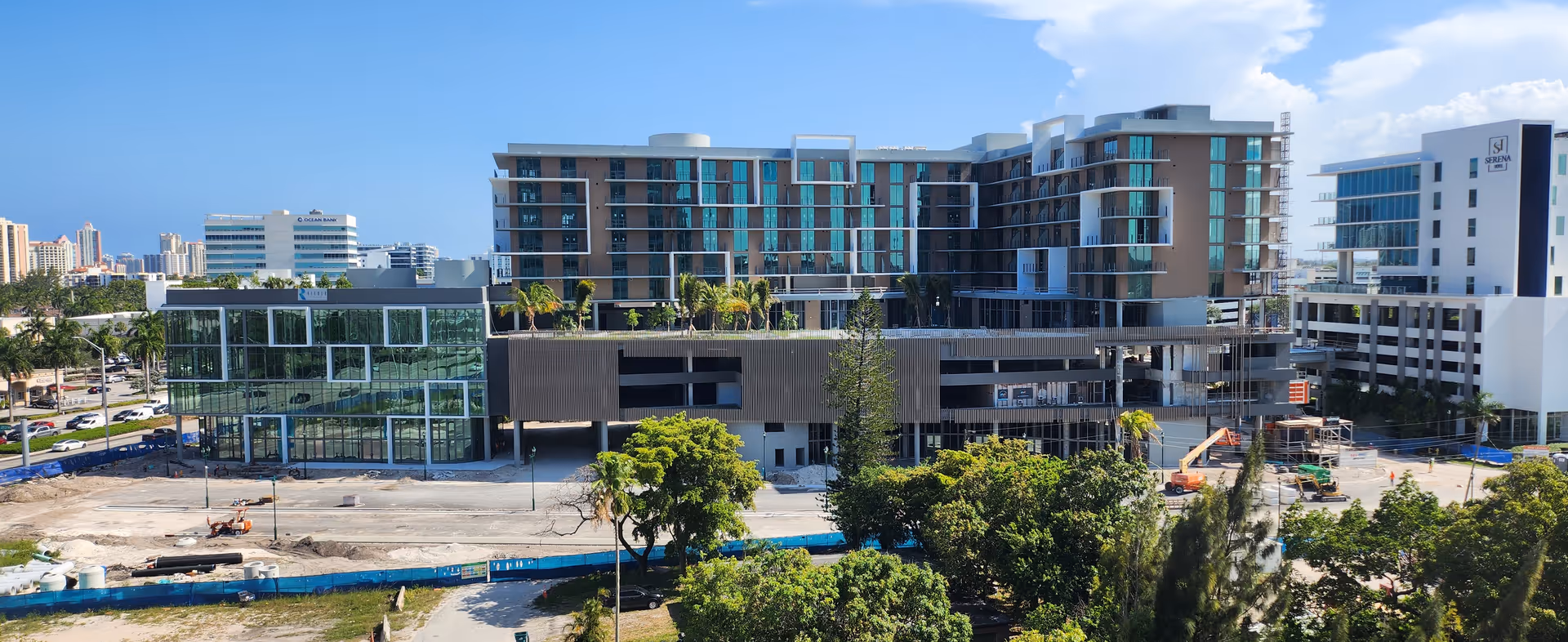 Exterior view of a modern multi-story senior living facility under construction with glass windows and balconies, surrounded by trees and other buildings under a clear blue sky.