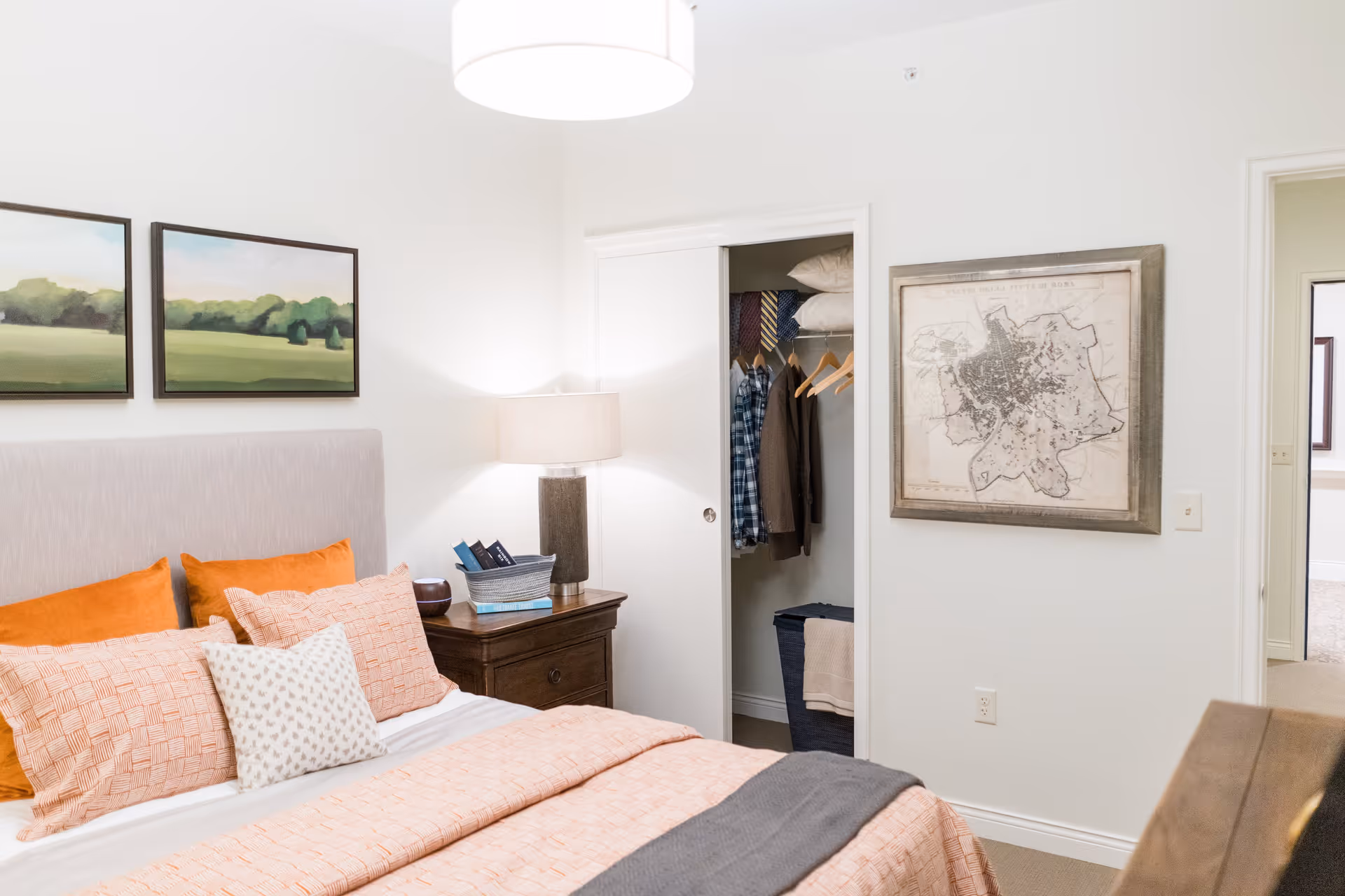 A cozy bedroom with a bed made up with orange and peach bedding and multiple pillows. There is a wooden nightstand with a lamp and some books beside the bed. On the wall above the bed are two landscape paintings. A closet with sliding doors is open, showing hanging clothes and pillows. A framed map hangs on the adjacent wall, and a doorway leads to another room.