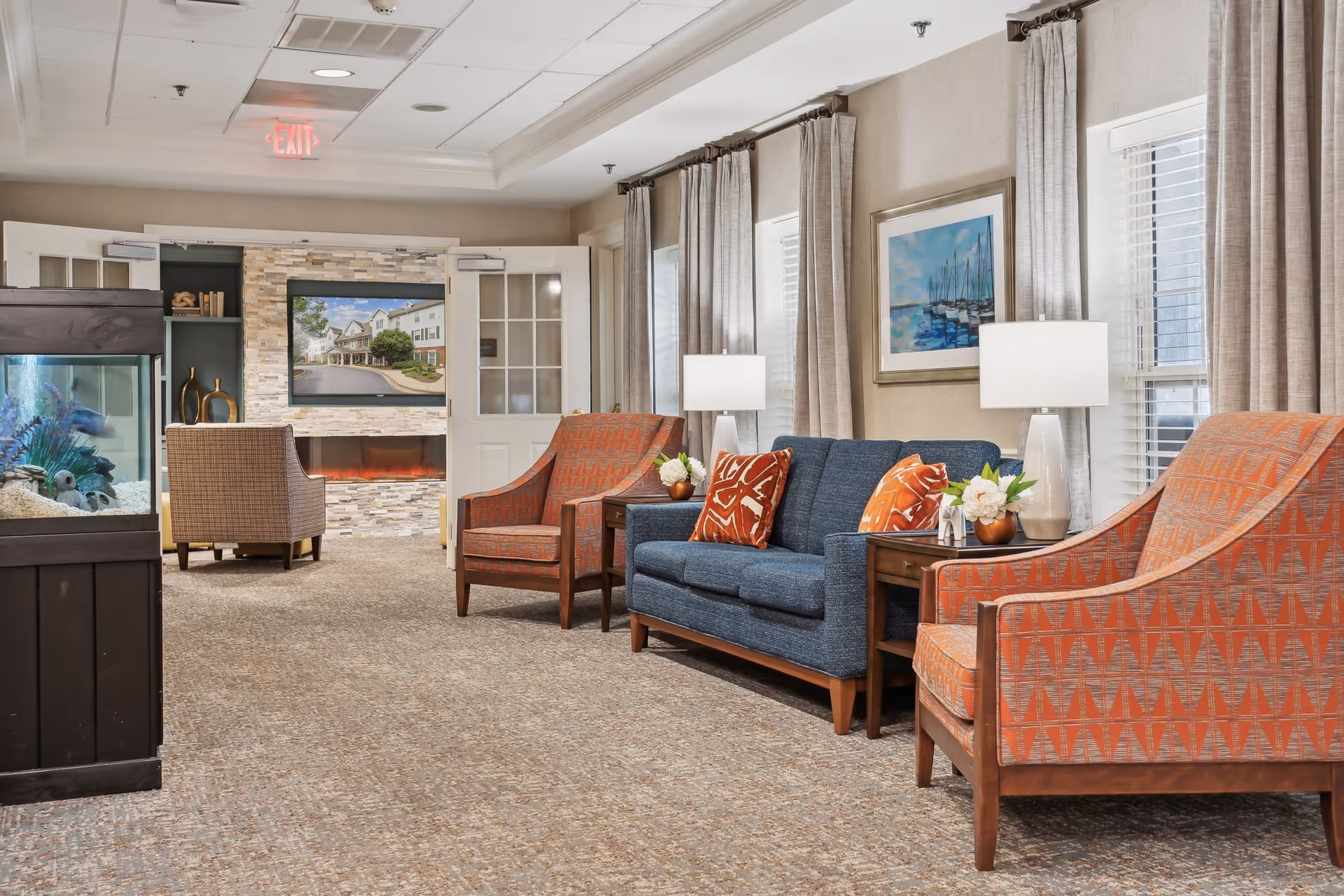 A cozy living room area in a senior living facility featuring a blue sofa with orange patterned pillows, two orange patterned armchairs, side tables with lamps and flower arrangements, a large fish tank on the left, and a wall-mounted TV above a modern electric fireplace. The room has beige walls, carpeted floor, large windows with curtains, and a framed painting of boats on the wall.