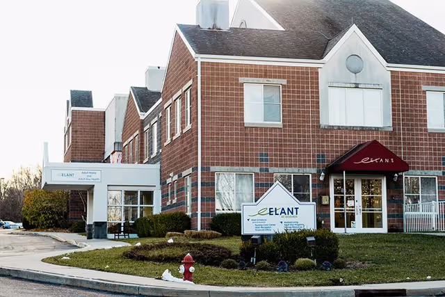 Exterior view of a multi-story brick building with a peaked roof and multiple windows. The building has a covered entrance with a sign that reads 'Elant at Goshen' and a red awning above the door with the word 'Elant'. There is a landscaped area with bushes and a fire hydrant near the curb in front of the building.