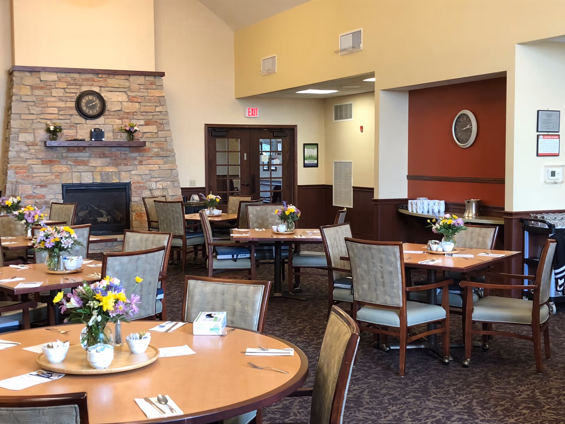 Dining room with multiple wooden tables and chairs arranged neatly. Each table has a vase with colorful flowers and place settings including napkins, utensils, and small bowls. A stone fireplace with a clock above it is visible on the left side of the room. The walls are painted in warm tones with a mix of beige and red accents. There are double glass doors and an exit sign in the background.