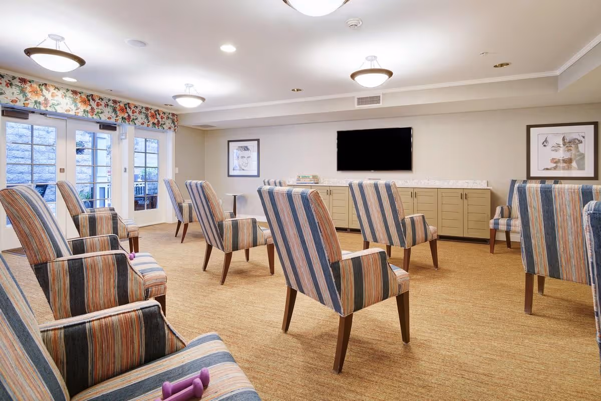 A well-lit common room with multiple striped upholstered chairs arranged in rows facing a wall-mounted flat-screen TV. The room has beige walls, carpeted floor, floral valance above glass doors leading outside, and framed artwork on the walls.