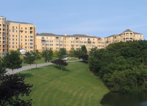 Exterior view of a multi-story senior living building set behind a landscaped lawn, trees, and a small pond.